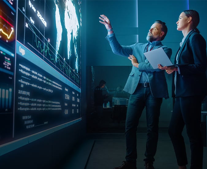 Two people gesturing to a map and data on a large video wall.